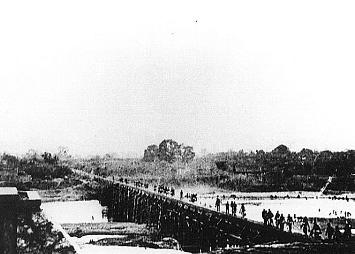 Widok na drewniany most z południowego brzegu rzeki Kwae Yai. Na moście widoczne postacie jeńców wojennych. W oddali widoczne zarysy przęseł mostu stalowo-betonowego.