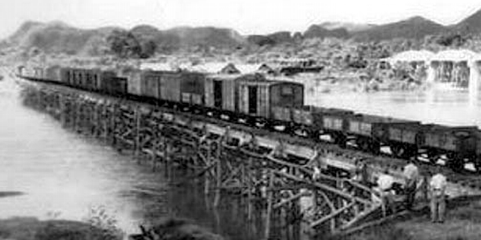 Pociąg towarowy przejeżdża przez most drewniany na rzece Kwai Yai. Zdjęcie wykonane przez japońskiego żołnierza ze wschodniego brzegu rzeki. W Prawym rogu fotografii widoczni żołnierze japońscy przyglądający się przejazdowi pociągu.